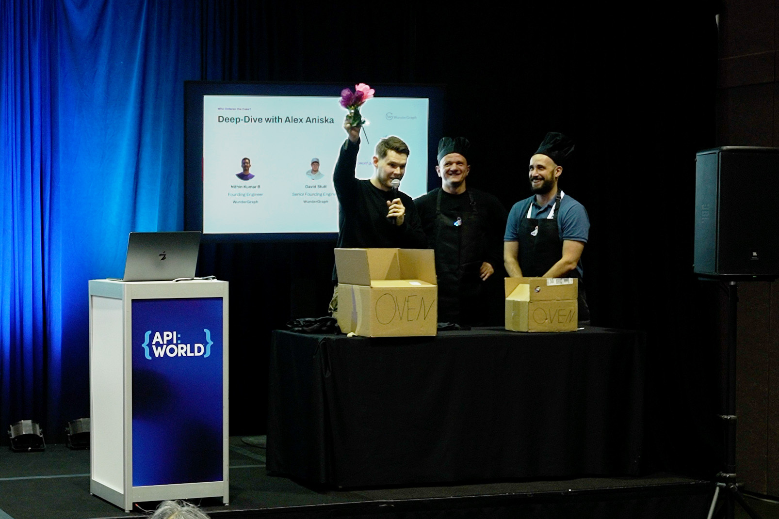 WunderGraph team on stage at API World in chef hats, with cardboard boxes labeled Oven, holding up a flower like a finished cake